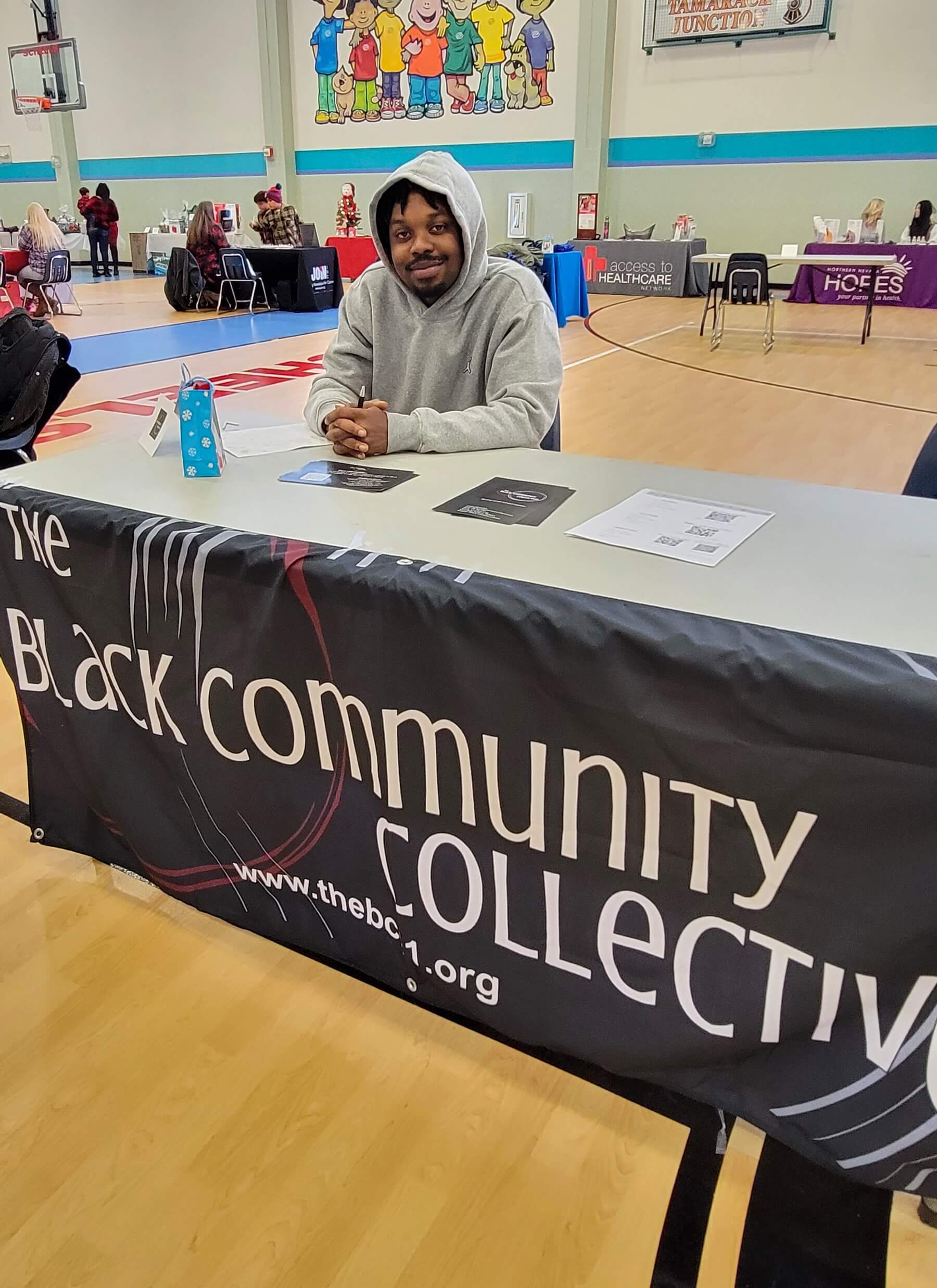 A man sitting at a table with a black community collective sign.
