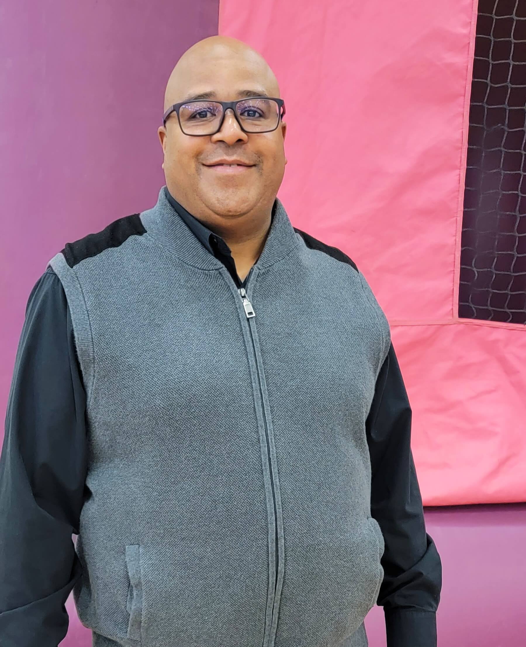 A man wearing glasses and a vest standing in front of a pink trampoline.