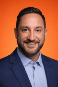 Professional headshot of a smiling man in a blue suit against an orange background.